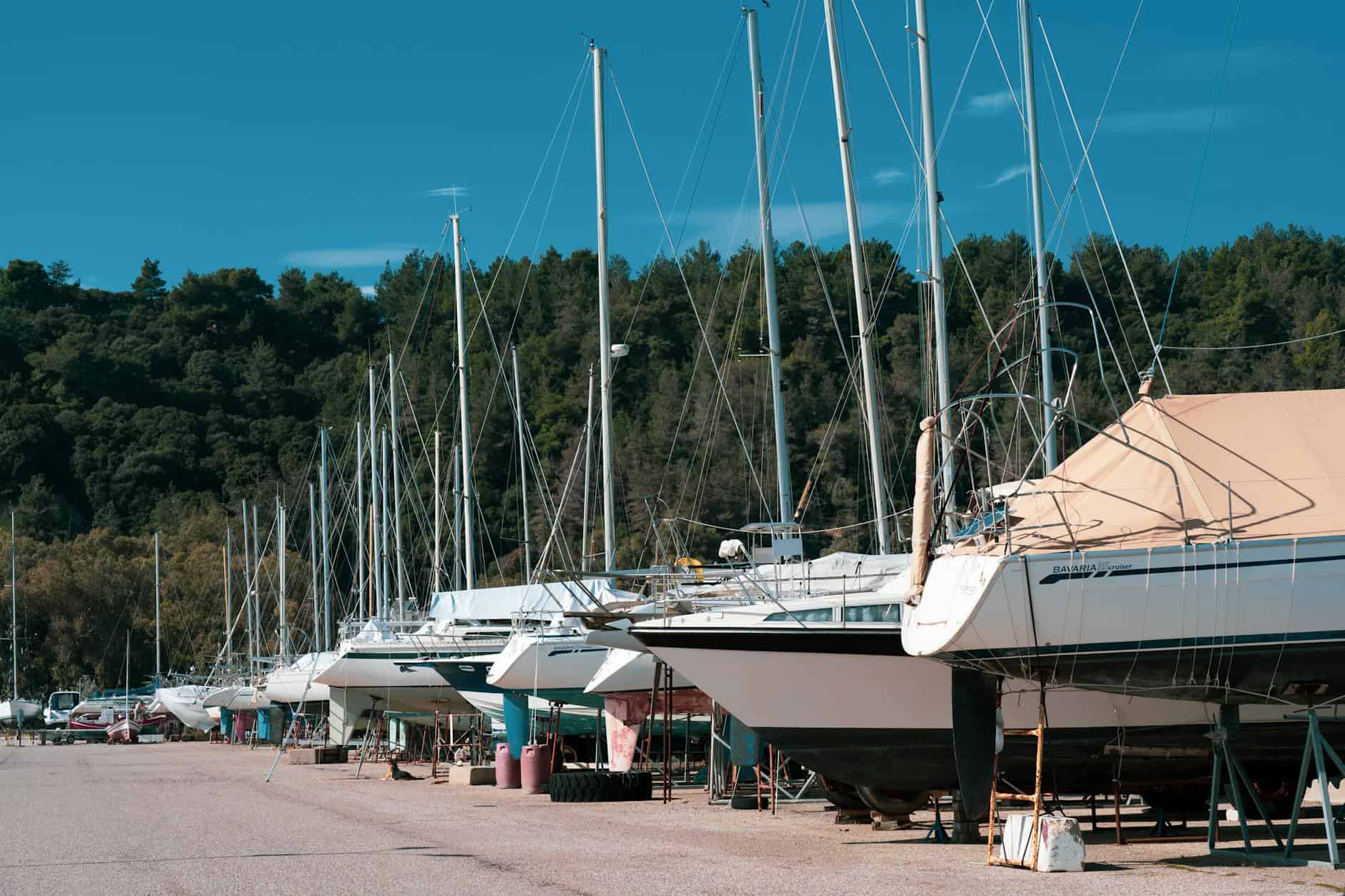 A row of sailboats docked on land, with masts reaching towards a clear blue sky and green trees in the background. Representing boats being stored. 