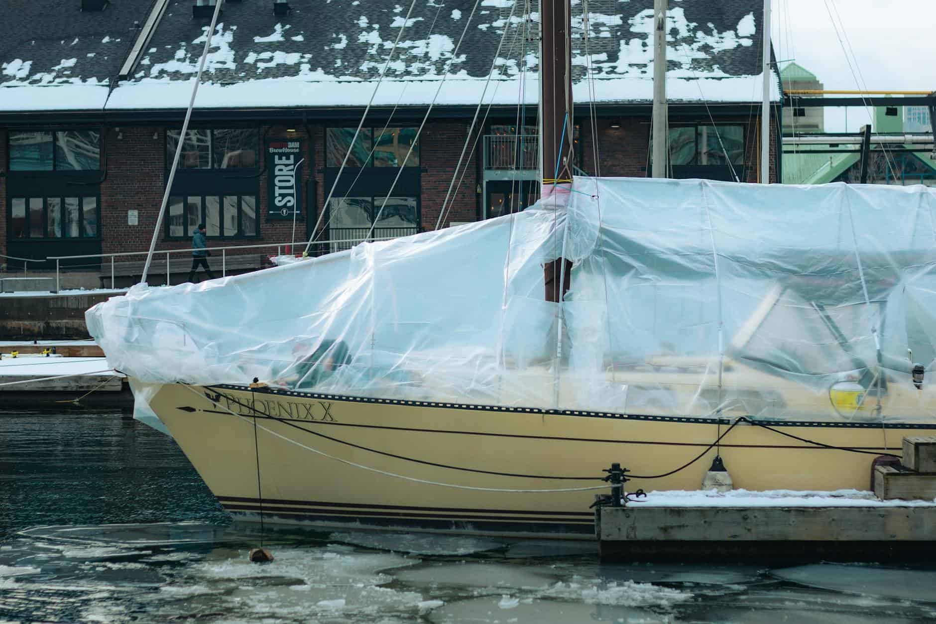 A boat covered with plastic tarps at a marina, surrounded by ice and snow, with a building in the background. representing winter boat storage. 
