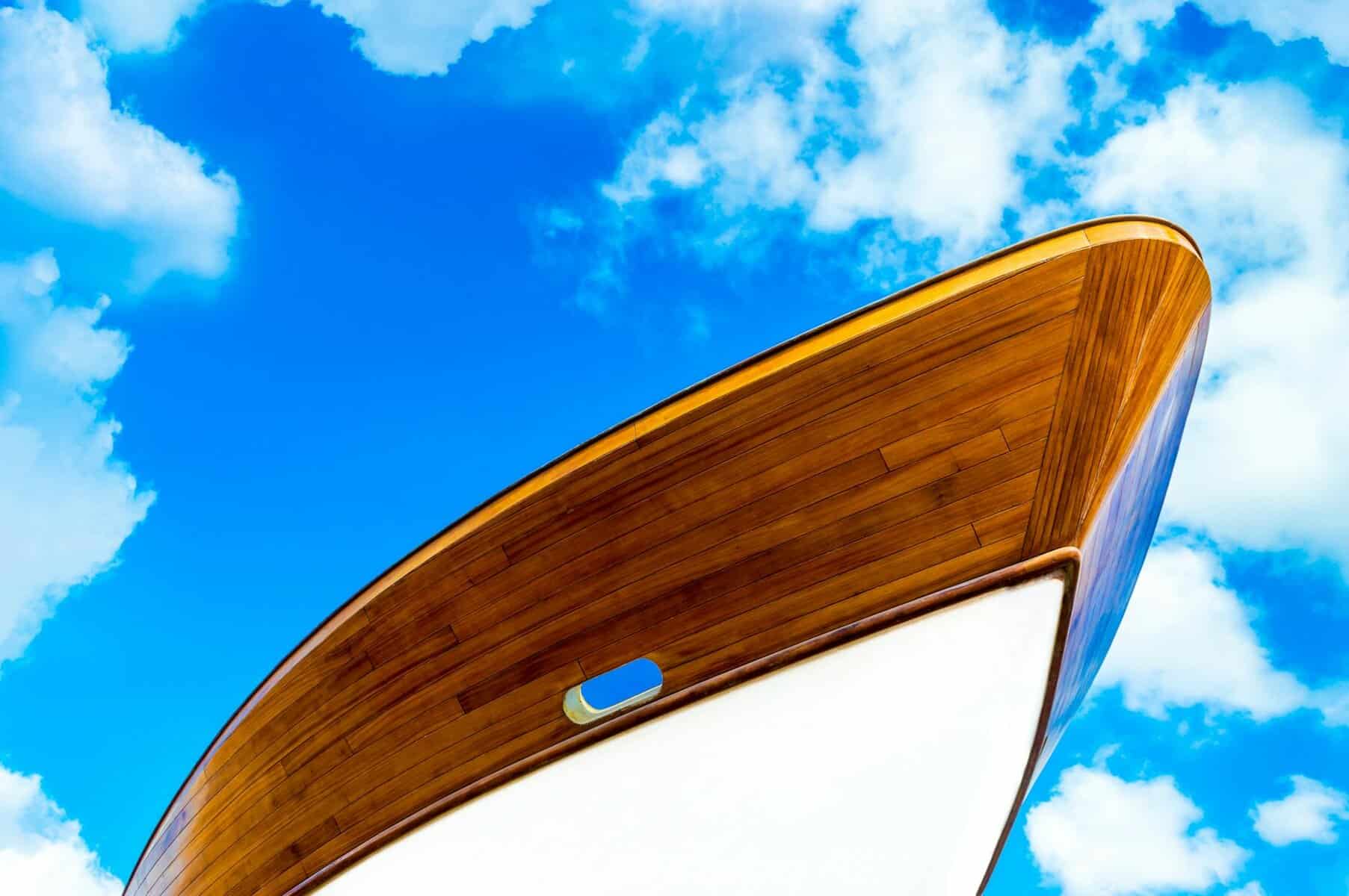 Varnished hull of a yacht with a blue and cloudy sky behind. 